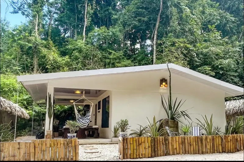 Single-storey white house with a covered porch, hanging hammock chair and bamboo fence, backed by dense forest