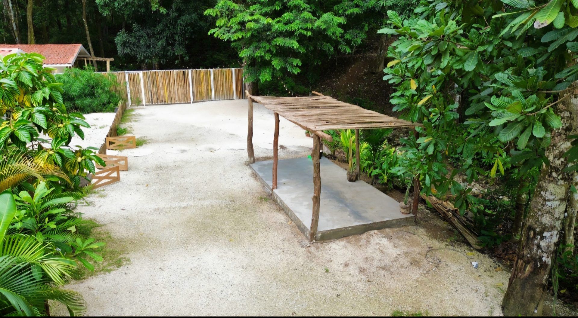 Gravel yard with a small rustic shade structure over a concrete slab, surrounded by tropical plants and a bamboo fence
