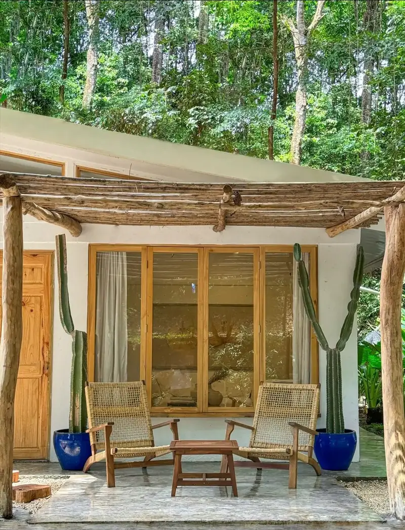 Covered front patio with two woven armchairs and a small table under a wooden pergola, backed by forest