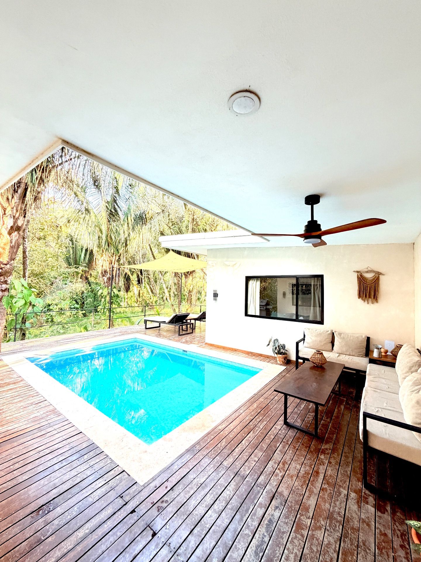 Covered wooden deck with ceiling fan and outdoor sofas beside a private pool, surrounded by palm trees