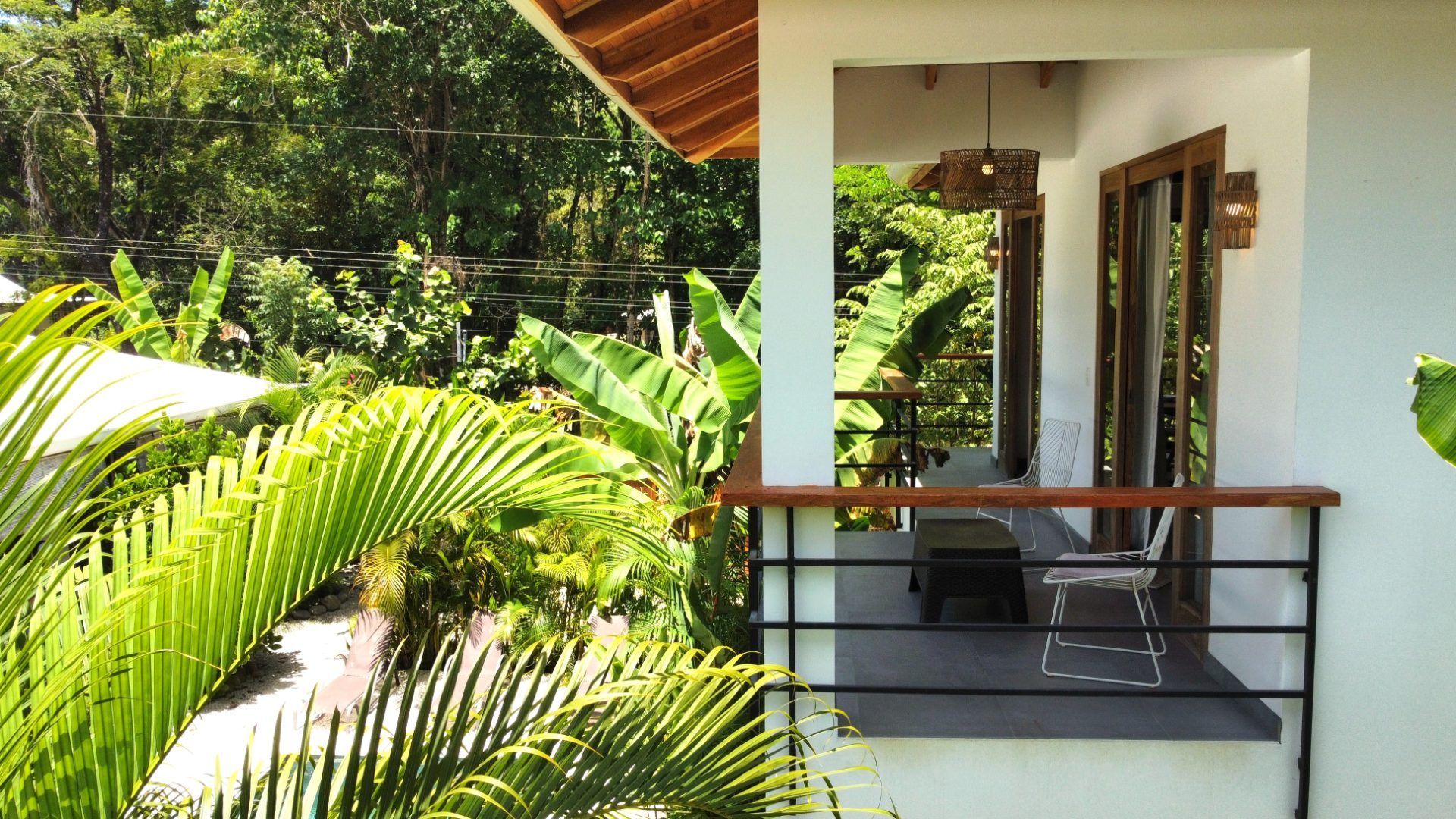 Covered upper-floor balcony with two wire chairs and a small table, surrounded by palms and tropical trees