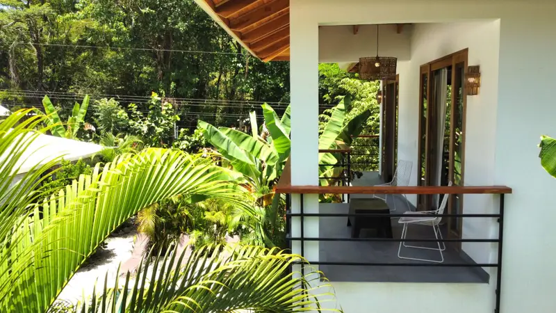 Covered upper-floor balcony with two wire chairs and a small table, surrounded by palms and tropical trees