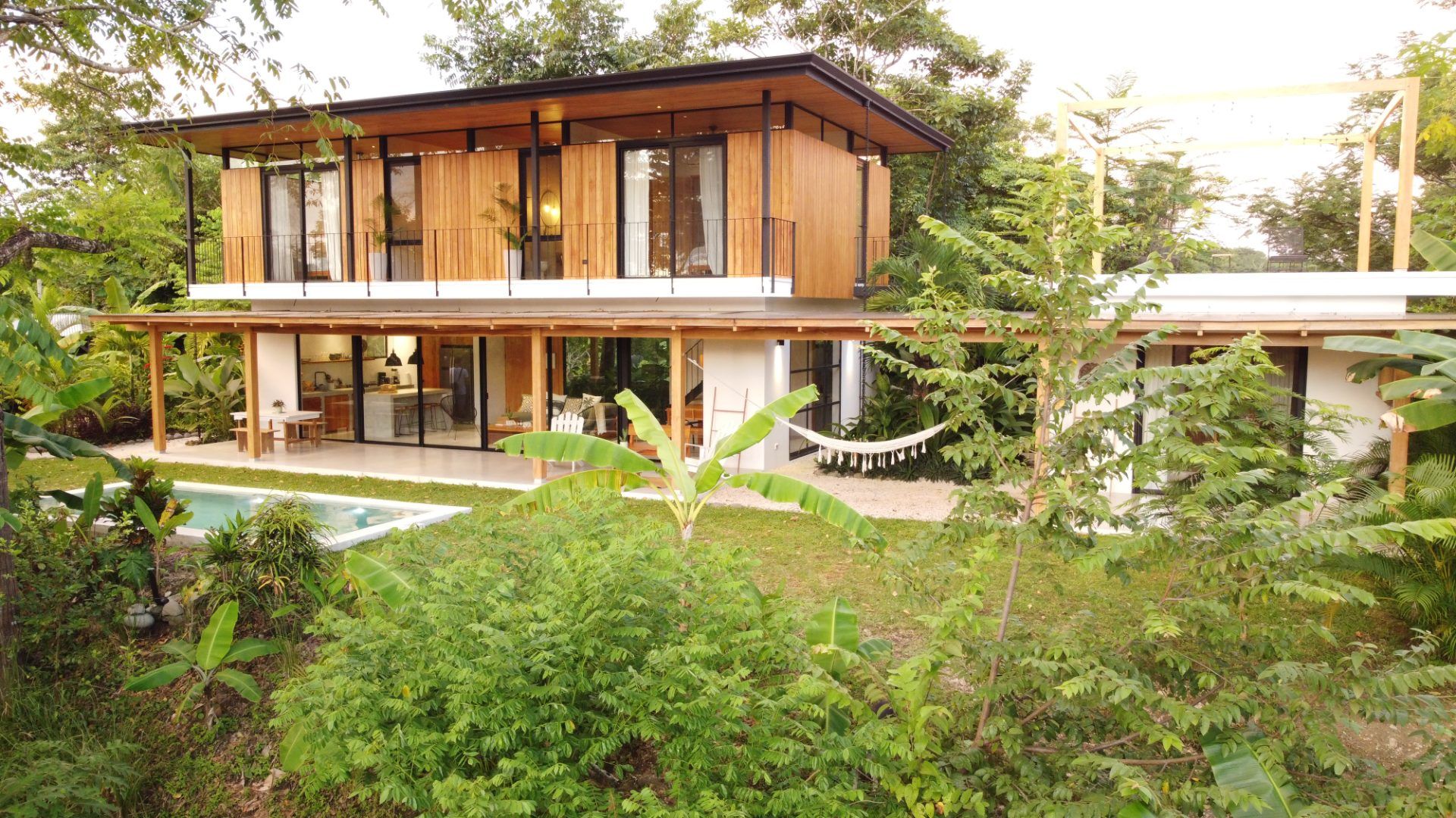 Two-storey villa with wood-clad upper floor and balcony, covered patio with hammock, and a pool in a tropical garden