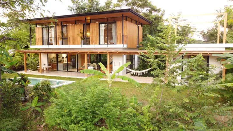 Two-storey villa with wood-clad upper floor and balcony, covered patio with hammock, and a pool in a tropical garden