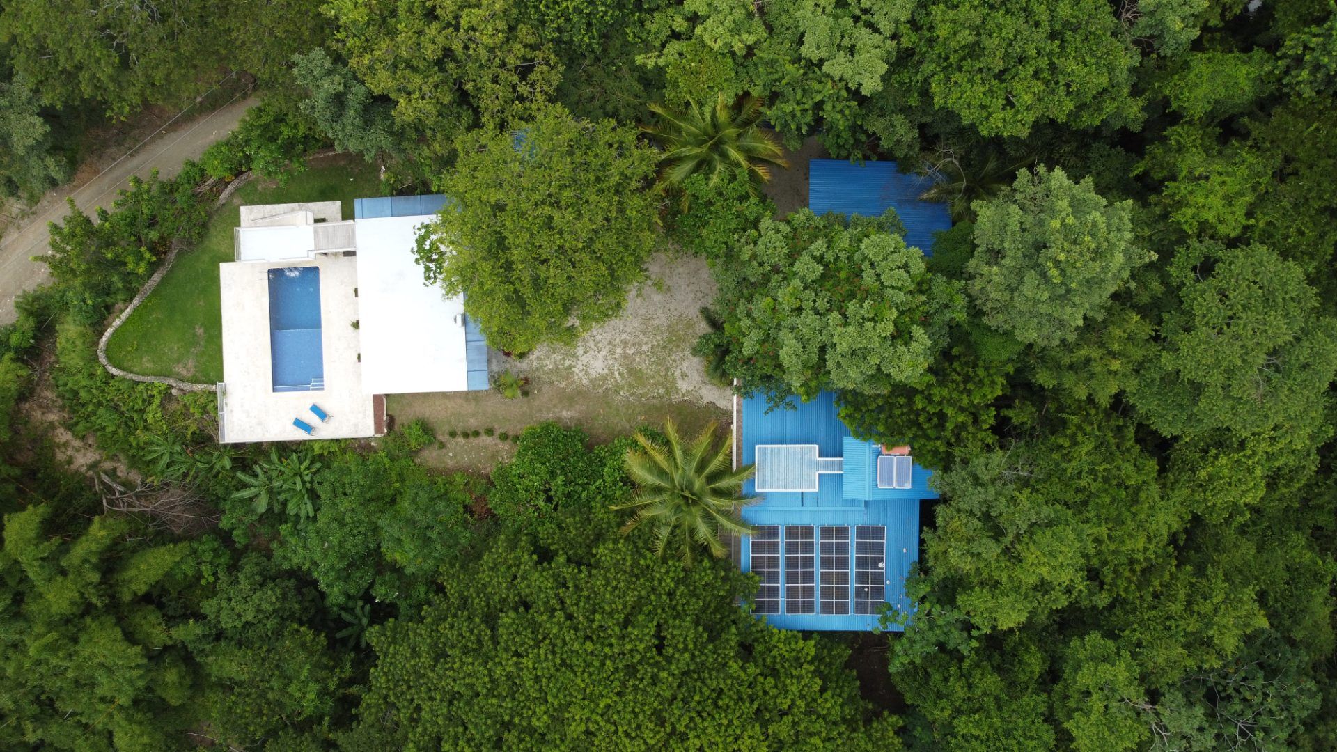 Aerial view of the compound: pool house with pool and lawn, and a blue-roofed house with solar panels among dense trees