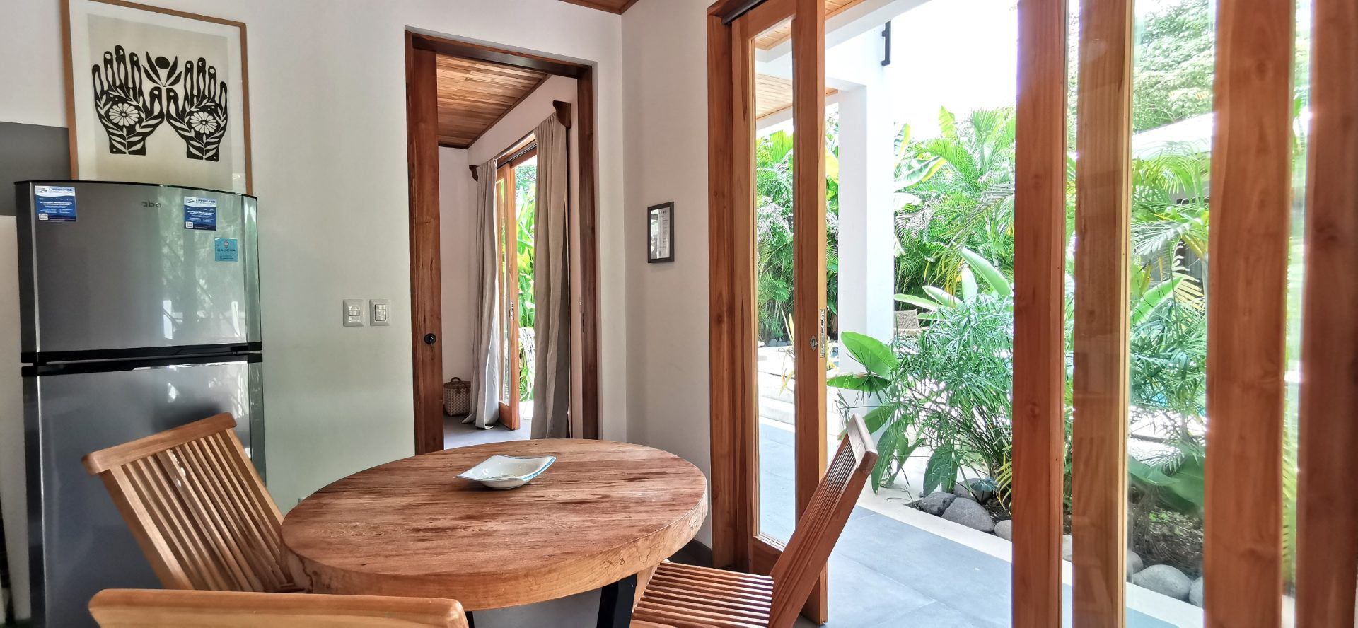 Round wooden dining table with two folding chairs beside a fridge, glass doors open to a tropical garden