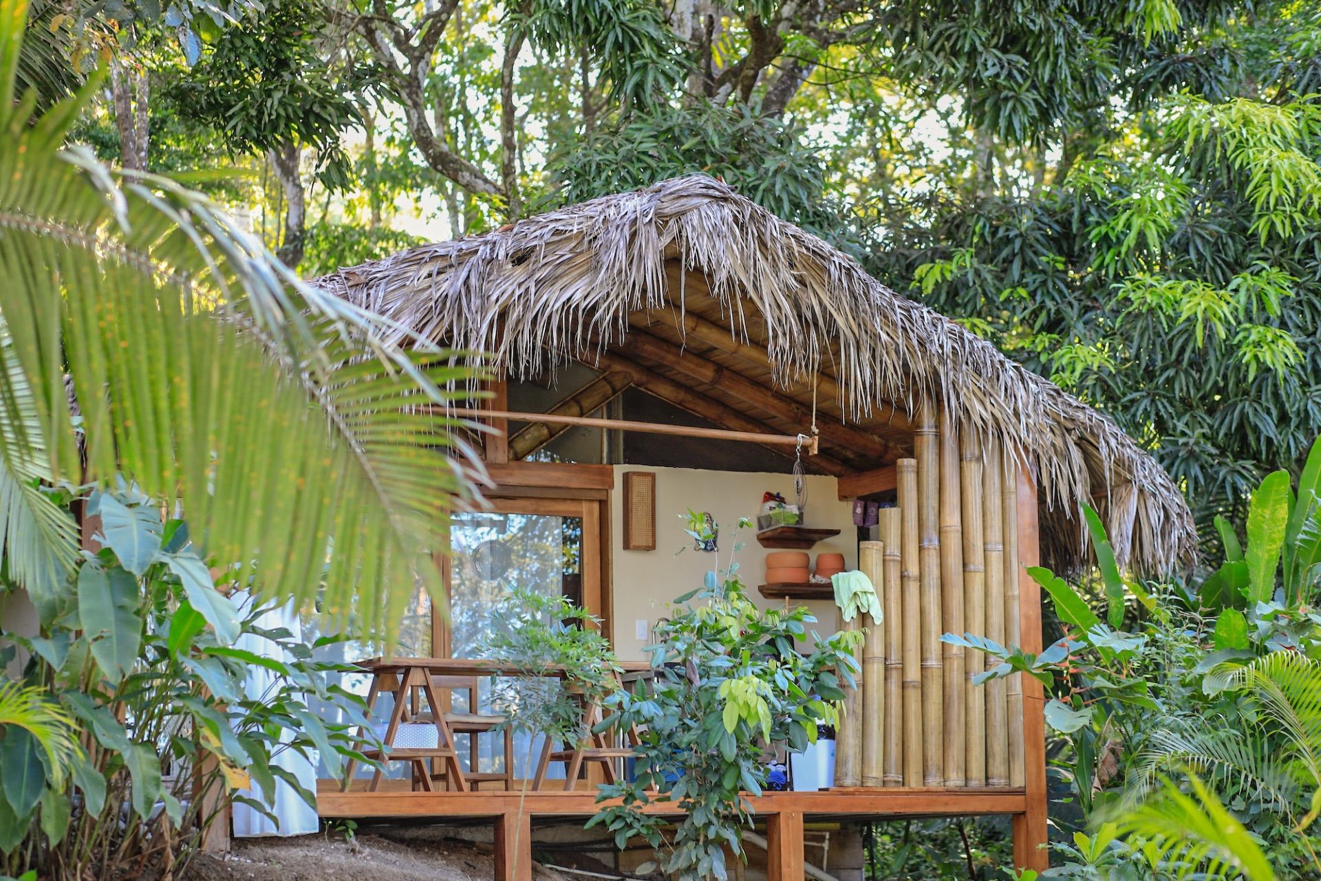Thatched-roof wooden bungalow on a raised deck surrounded by tropical plants and trees, with a table on the porch