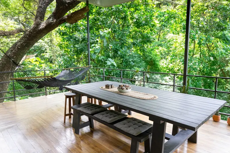 Covered wooden deck with a long dining table, cushioned bench and hammock, surrounded by dense trees