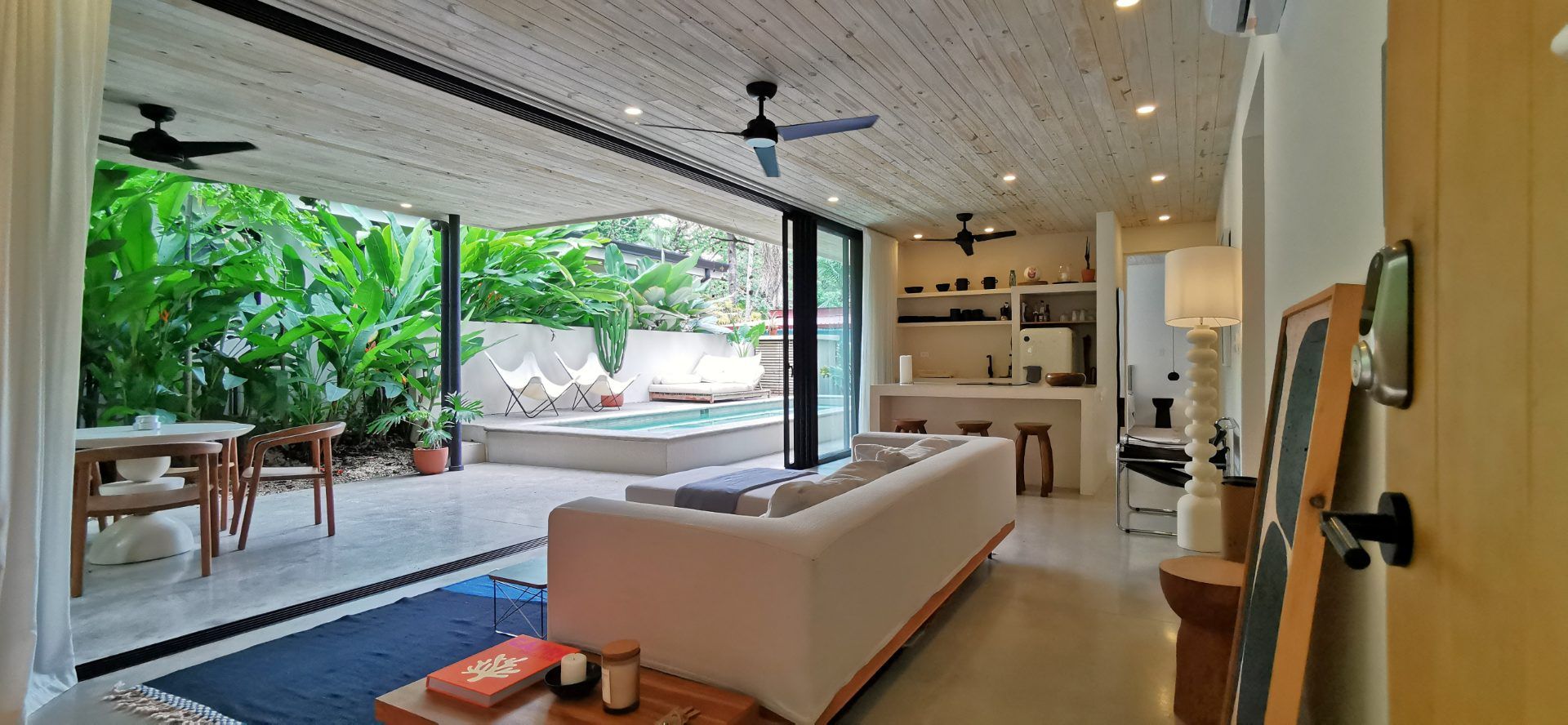 Open-plan living room with sofa, ceiling fans and A/C, opening onto a covered patio with dining table and private pool