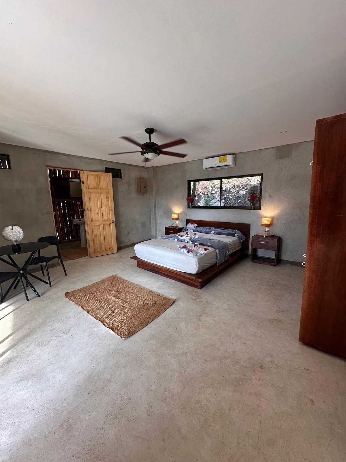Bedroom with a queen platform bed, ceiling fan, wall A/C unit, small dining table and door to the bathroom