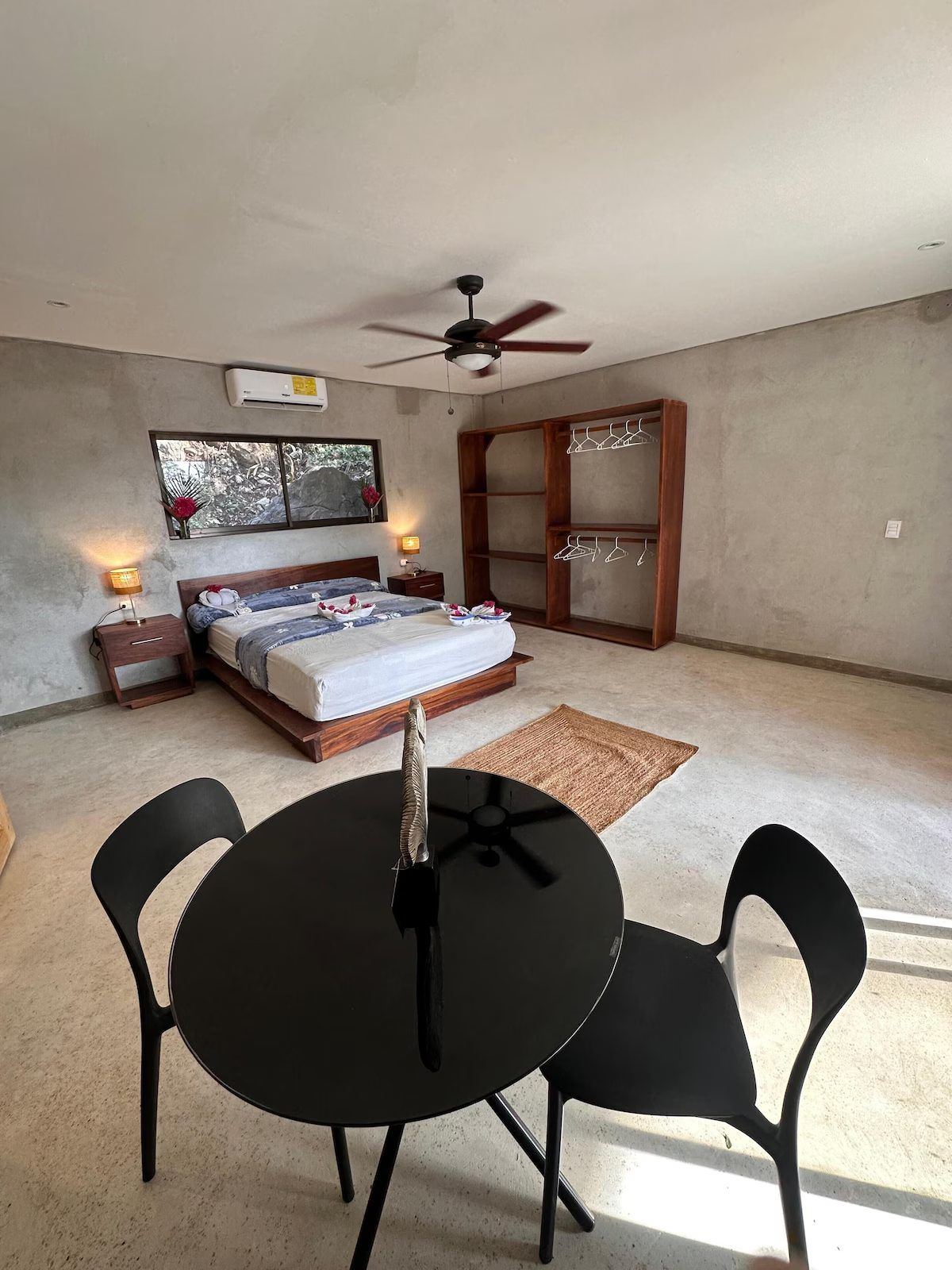 Studio with a queen bed, open wooden wardrobe, ceiling fan, wall A/C unit and a round table for two