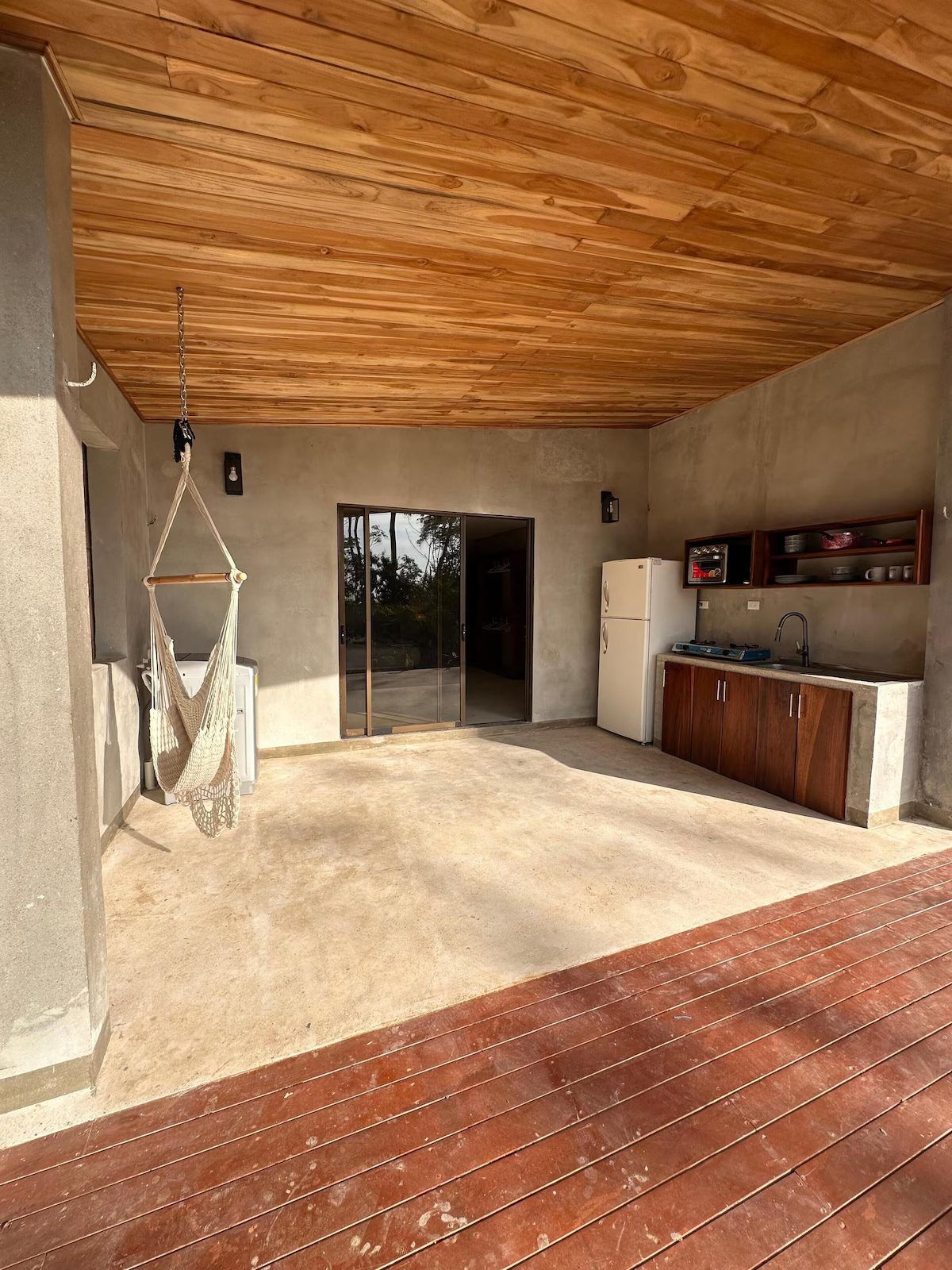 Covered terrace with a hanging chair, washing machine and outdoor kitchen with fridge and sink beside a sliding door