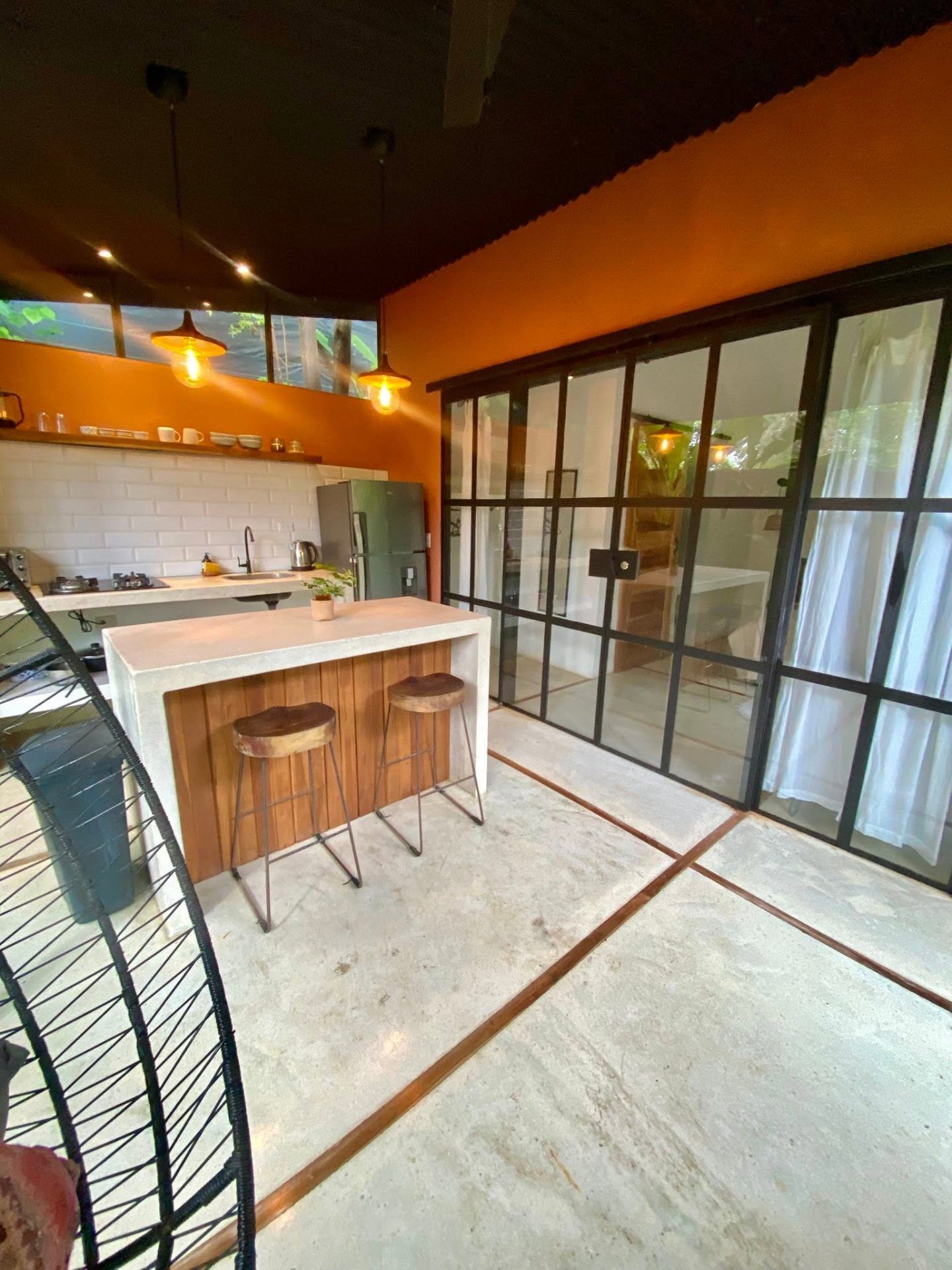 Covered open-air kitchen with gas hob, fridge and breakfast bar with two stools beside glass doors to the bedroom