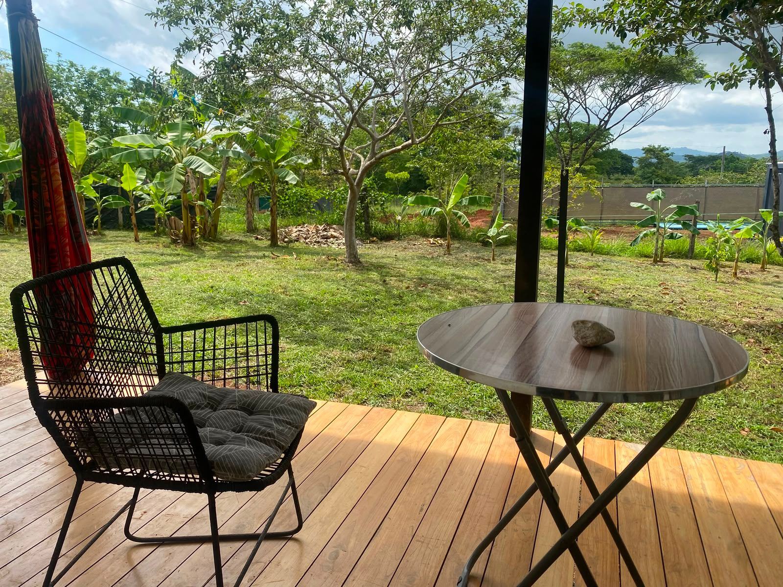 Wooden deck with a cushioned chair, small round table and hammock, facing a lawn with banana plants and trees