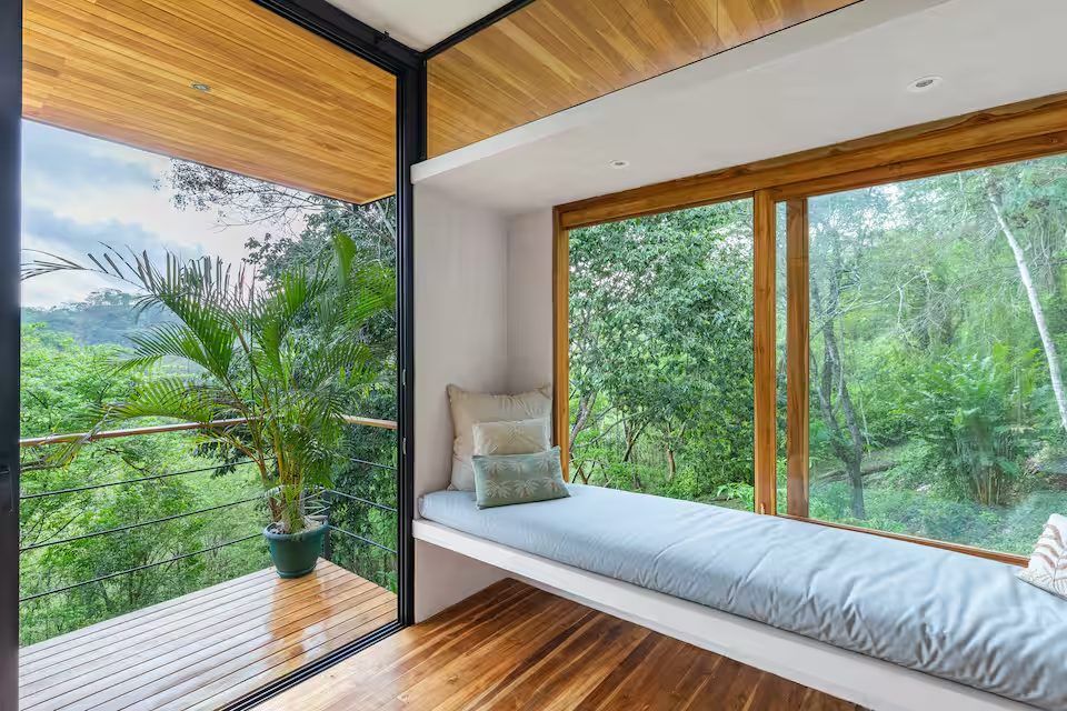Built-in window seat with cushions beside floor-to-ceiling glass opening onto a wooden balcony facing forest