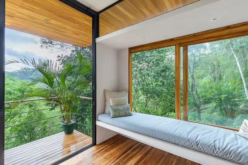 Built-in window seat with cushions beside floor-to-ceiling glass opening onto a wooden balcony facing forest