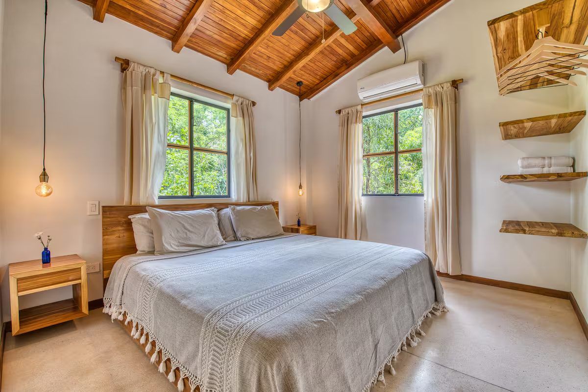 Bedroom with a king bed, wall-mounted A/C, ceiling fan and open shelving, with windows onto trees