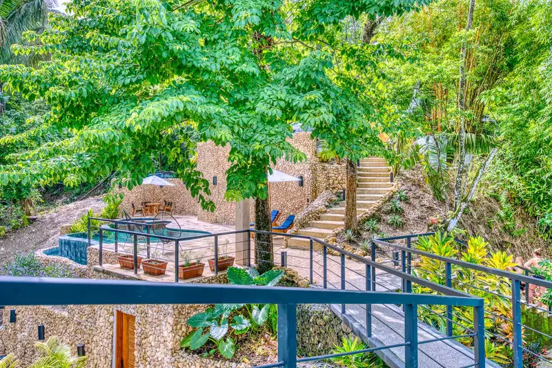 Stone-walled pool terrace with loungers and an umbrella-shaded dining table, reached by a railed walkway through trees
