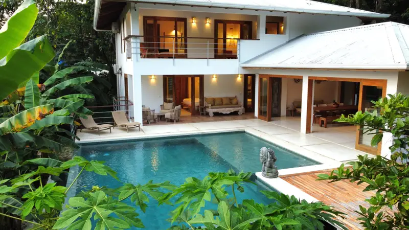 Two-storey white villa with a private pool, loungers, covered patio seating and an upper balcony, surrounded by tropical plants