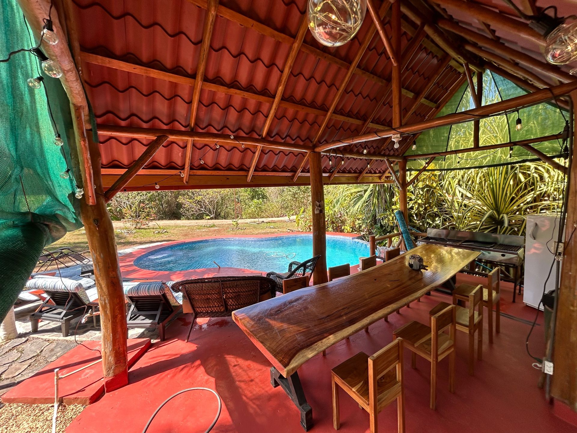 Covered outdoor dining area with a long wooden table, a large grill and a fridge, beside a kidney-shaped pool