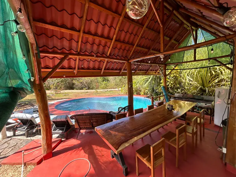 Covered outdoor dining area with a long wooden table, a large grill and a fridge, beside a kidney-shaped pool