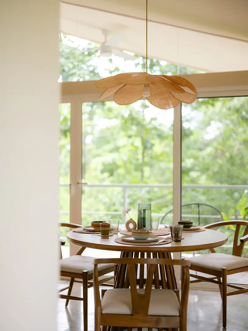 Round wooden dining table set for four under a rattan pendant, with glass doors to a balcony and trees