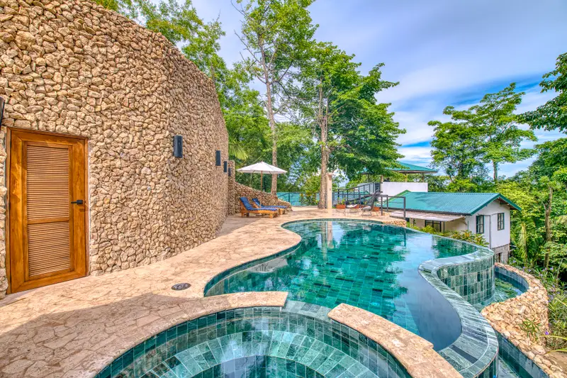 Tiled infinity pool with an attached spa pool, stone deck and loungers under an umbrella, surrounded by trees