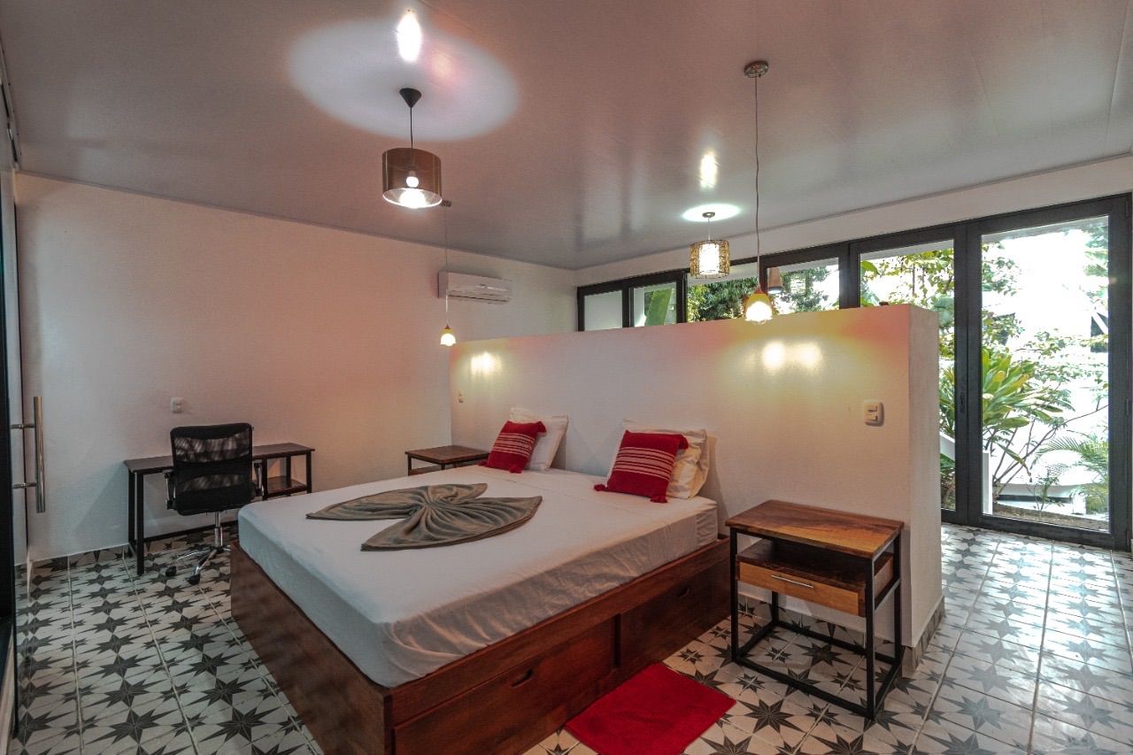 Bedroom with a queen bed, wall-mounted A/C, a desk and chair, and glass doors looking onto plants