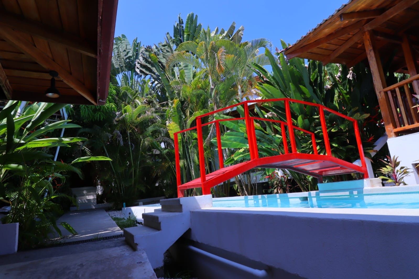 Pool with a red footbridge over it, surrounded by tropical plants and wooden buildings