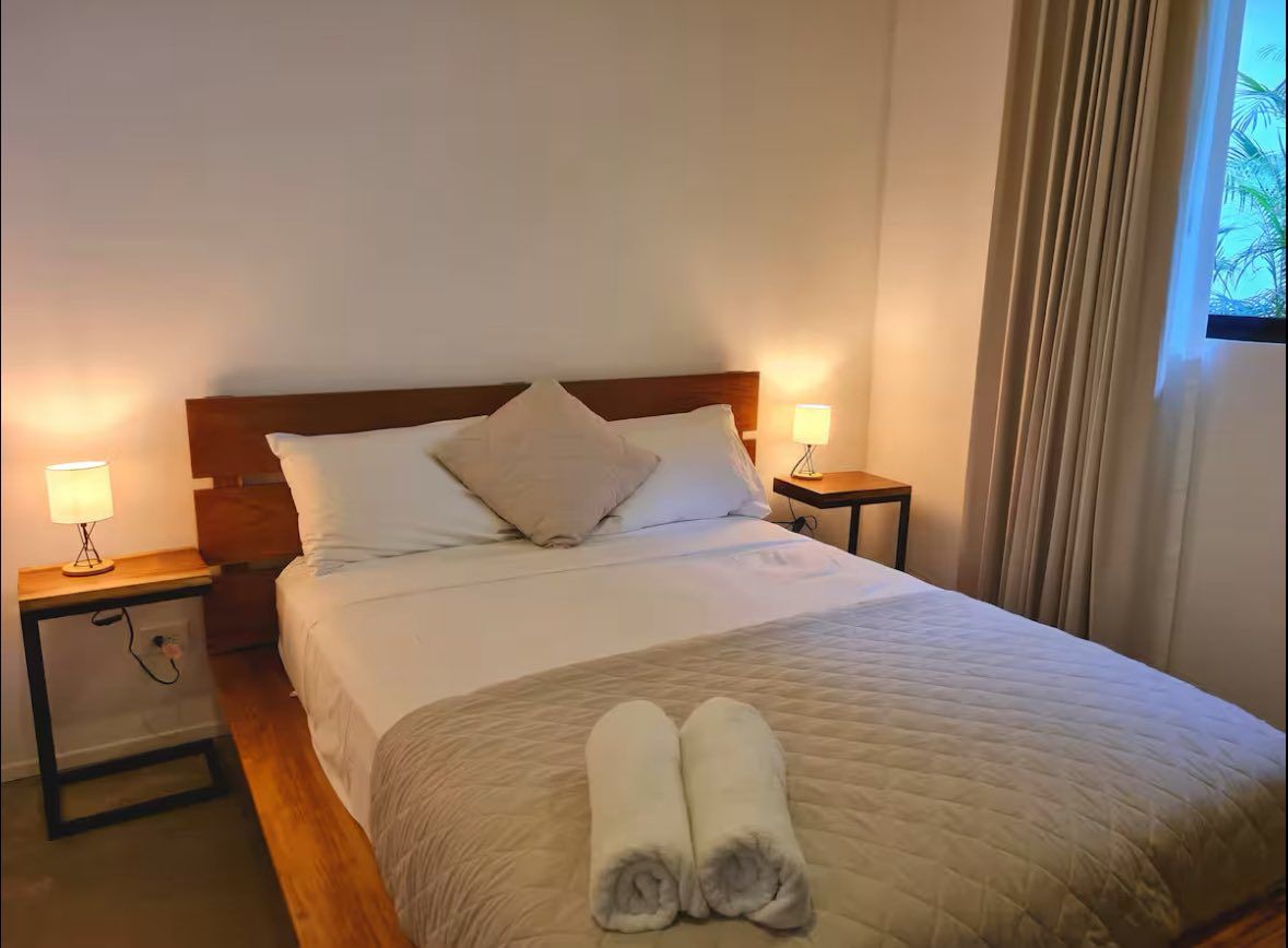 Bedroom with a queen bed, wooden headboard, two bedside lamps and a curtained window onto greenery