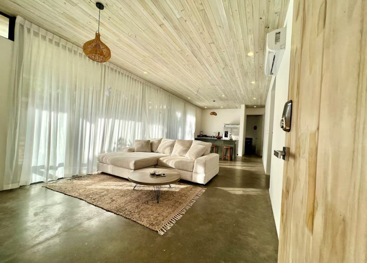 Open living room with a sectional sofa, wall-mounted A/C and kitchen counter with stools at the back