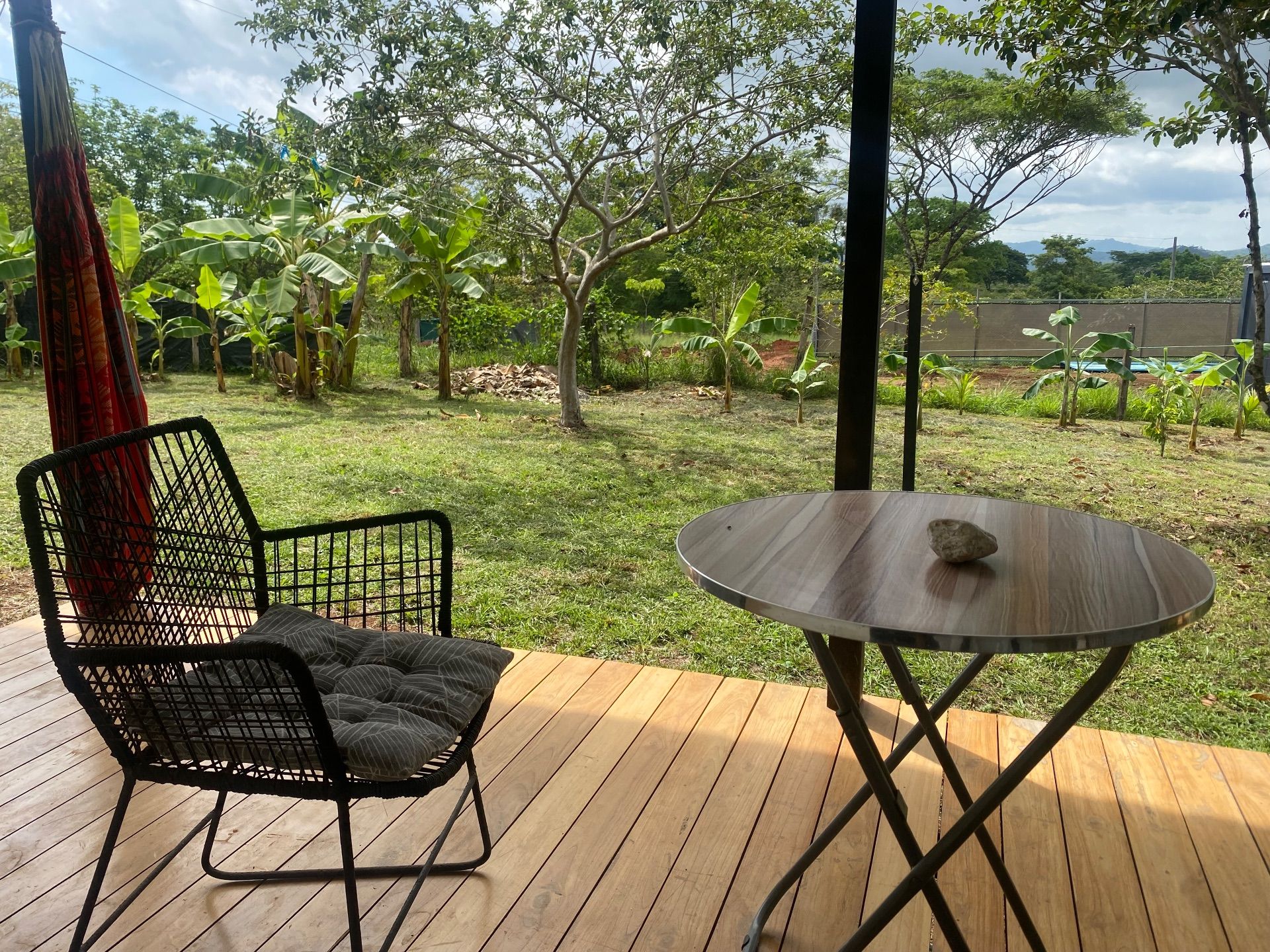 Wooden deck with a cushioned chair, small round table and hammock, facing a lawn with banana plants and trees