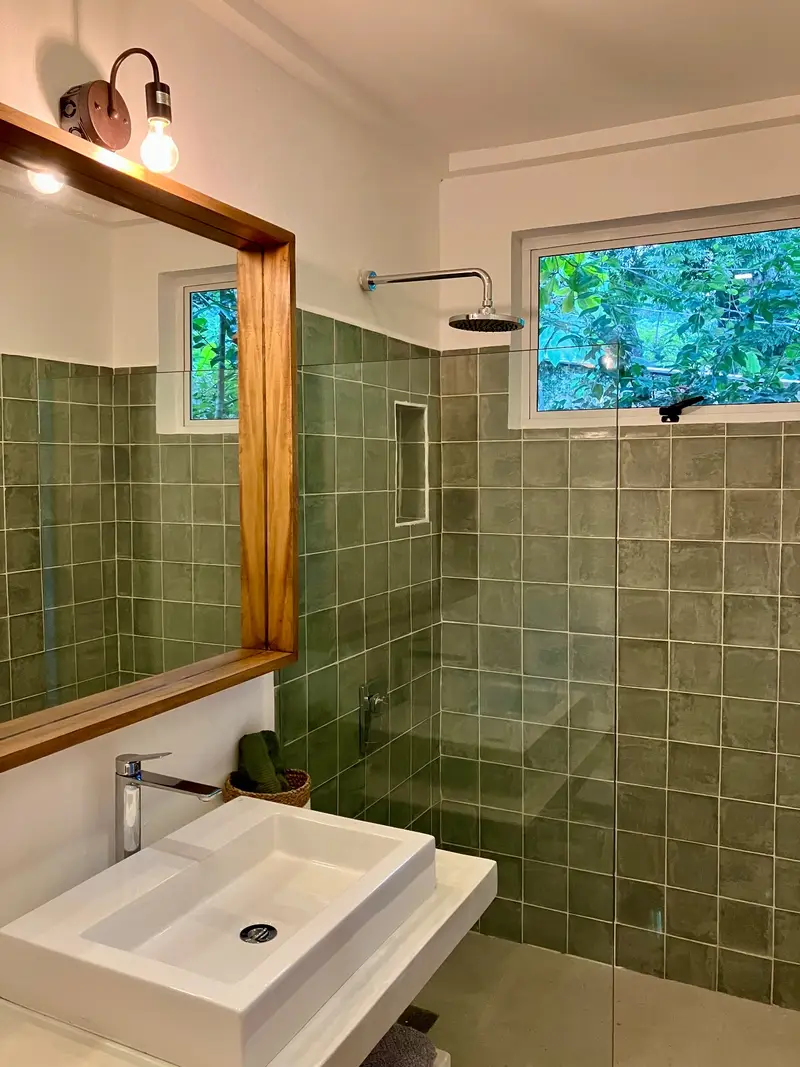 Bathroom with green-tiled walk-in rain shower behind glass, vessel sink and a window onto trees