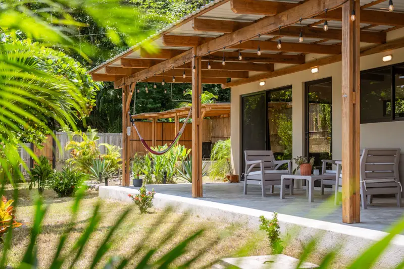 Covered patio with lounge chairs, a hammock and string lights, facing a fenced tropical garden